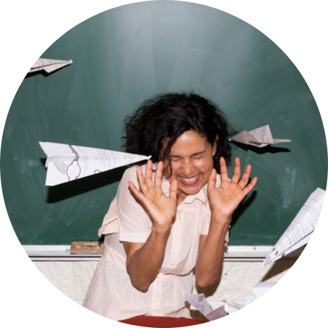 A woman with dark hair dodging paper airplanes thrown at her in front of a green chalkboard.